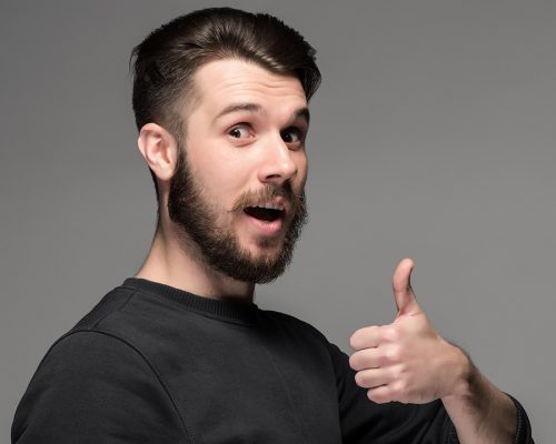 Closeup portrait of handsome young smiling man, giving a raised finger on gray background. Positive human emotions